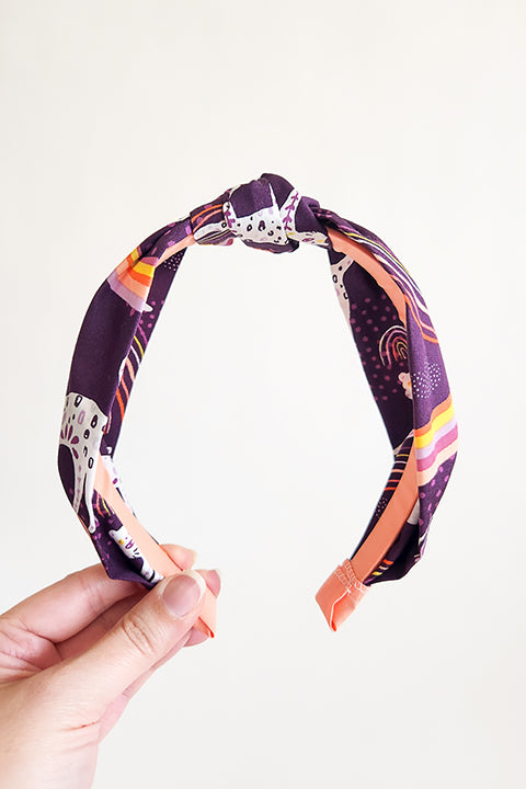 image of a hand holding a top knot styled handmade headband in front of a white background. Sustainably made from 100% cotton fabric scraps the print features boho white jaguars and yellow, pink and coral rainbows on a dark purple background.
