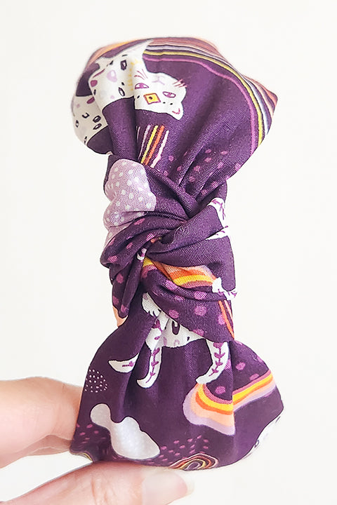 image of a hand holding a top knot styled handmade headband in front of a white background. Sustainably made from 100% cotton fabric scraps the print features boho white jaguars and yellow, pink and coral rainbows on a dark purple background.