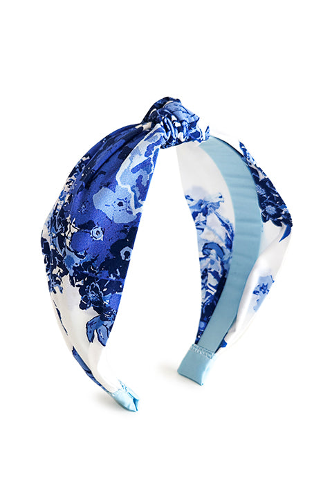 image of a handmade top knot style headband in front of a white background. The headband is made from upcycled 100% cotton fabric scraps in a blue floral rose print on a white background.