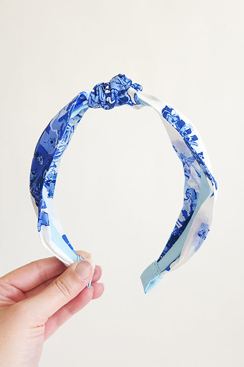 image of a hand holding a handmade top knot style headband in front of a white background. The headband is made from upcycled 100% cotton fabric scraps in a blue floral rose print on a white background.