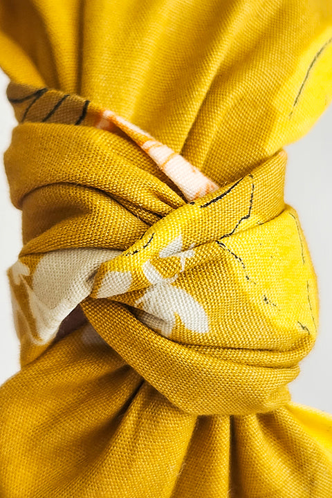 very close up detailed view of the twisted top knot on a top knot style sustainable handmade headband in front of a white background.  The 100% cotton material was upcycled from scraps and features a springy floral print in blush pinks on a mustard yellow background.