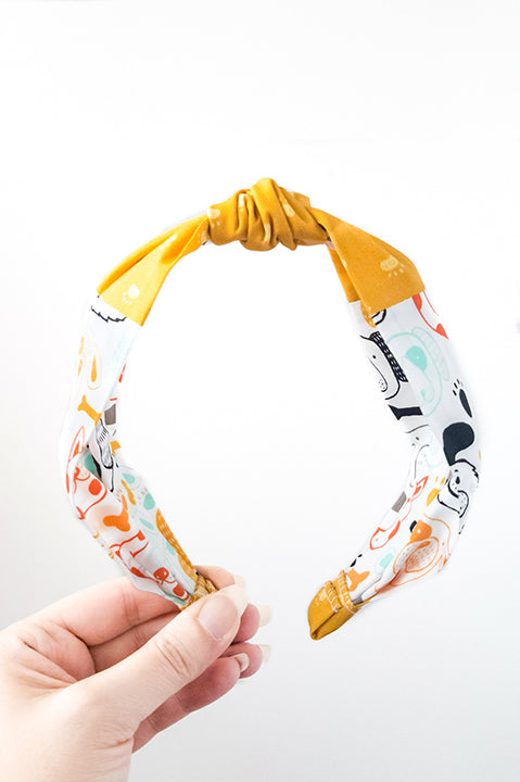front view of an upcycled cotton fabric headband featuring a top knot and two fabric prints, one featuring mustard yellow paw prints and the other with colorful dog faces