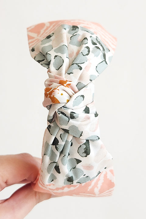 image of a hand holding a two tone top knot style handmade headband. Sustainably made from 100% cotton fabric scraps in two prints, one a pink and sage green watercolor dot floral and the other white ferns on a blush pink background.