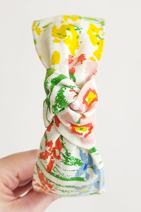 image of a hand holding a handmade top knot style headband in front of a white background. Sustainably made from 100% cotton vintage floral curtains. the floral print is in red, blue, yellow, green and white.