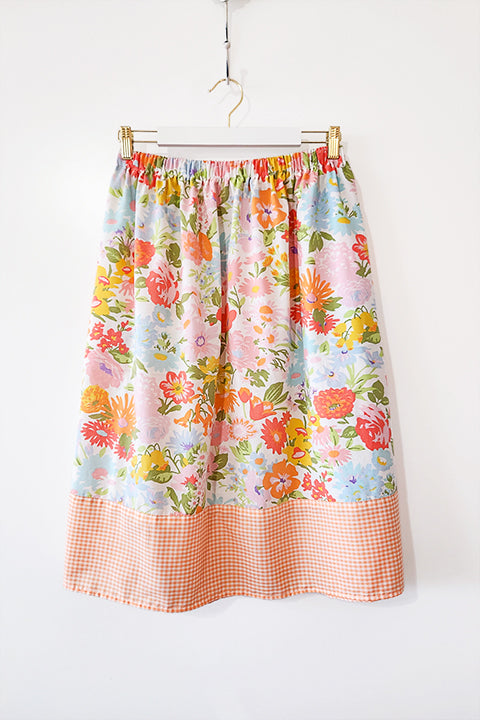 image of an elastic waist gathered skirt hanging on a wall. the skirt is made from vintage materials, a colorful floral print paired with a small, gingham check border in peach and white